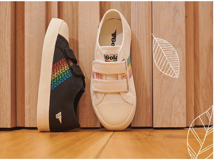 Black and white sneakers with rainbow stripes and velcro straps, leaning against a wooden wall.