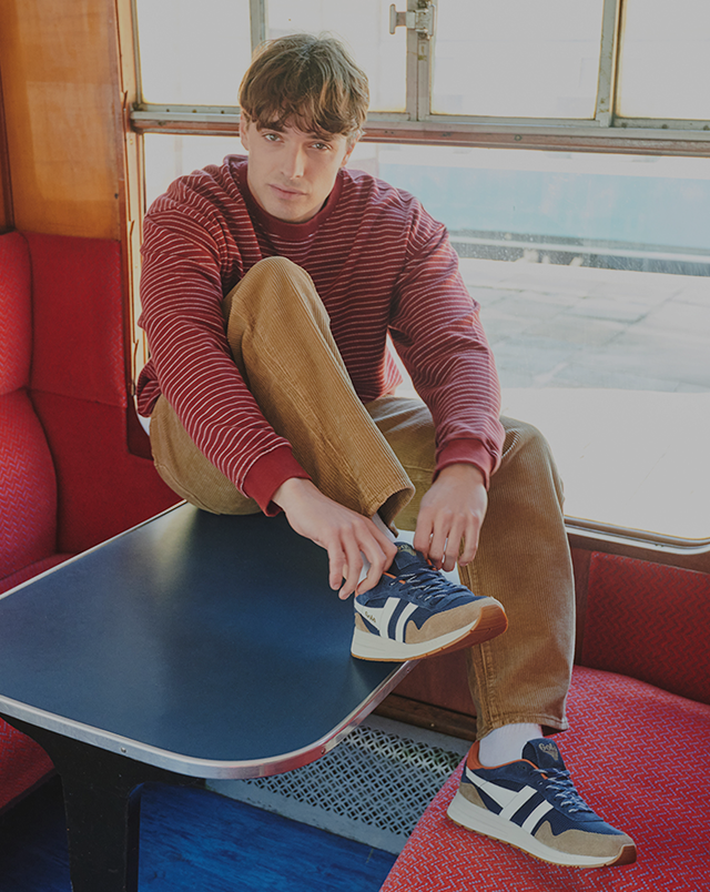Man in a striped shirt and corduroy pants tying his navy and white sneakers on a train.
