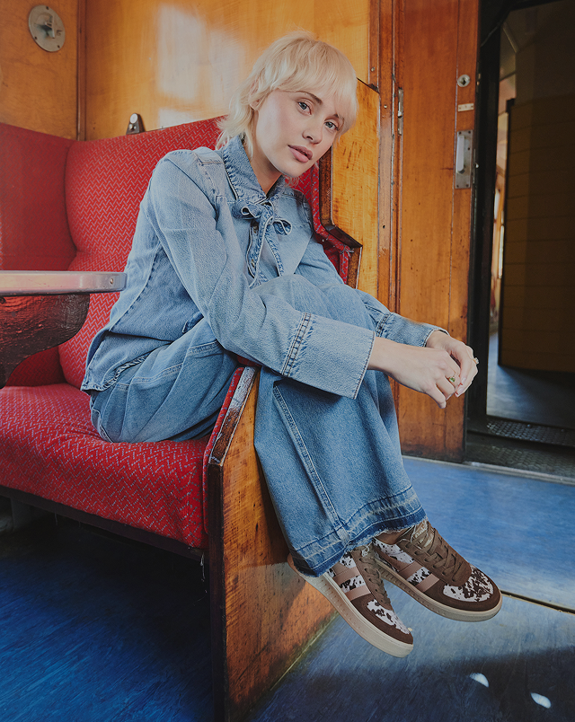 A woman with short blonde hair sits on a red train seat, wearing a denim jacket, wide-leg jeans, and brown sneakers.